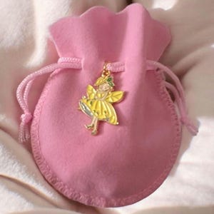 May include: A small, soft pink velvet drawstring pouch featuring a gold-toned metal charm shaped like a fairy with yellow wings and a yellow dress. The charm is attached to the drawstring cord of the pouch, which is made of a plush fabric.