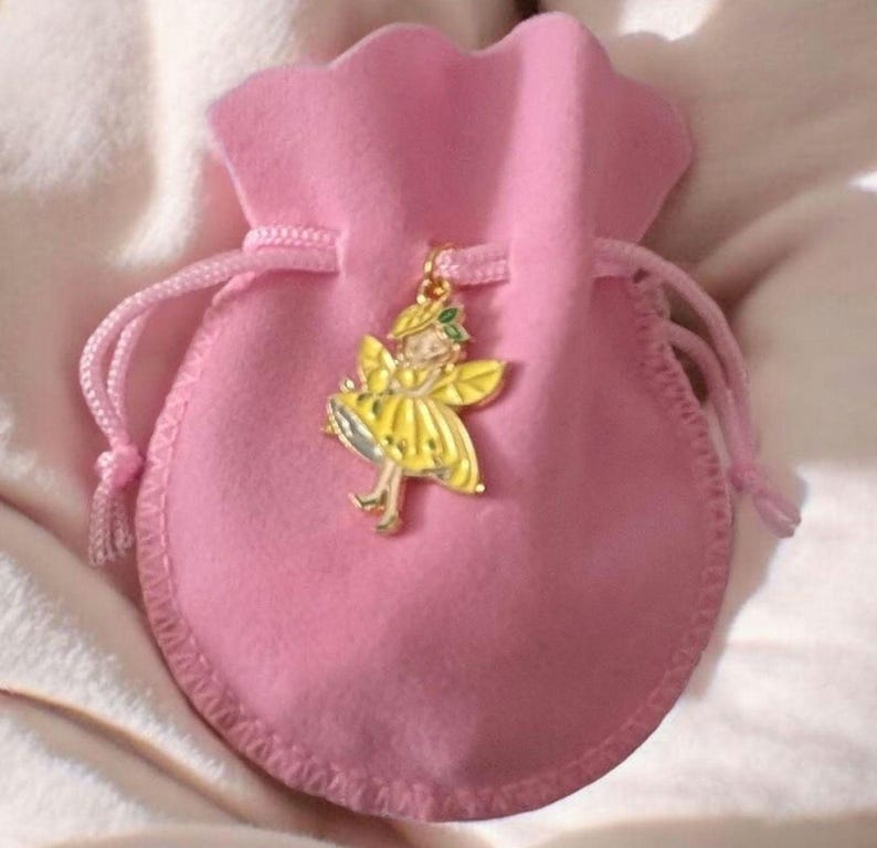 May include: A small, soft pink velvet drawstring pouch featuring a gold-toned metal charm shaped like a fairy with yellow wings and a yellow dress. The charm is attached to the drawstring cord of the pouch, which is made of a plush fabric.