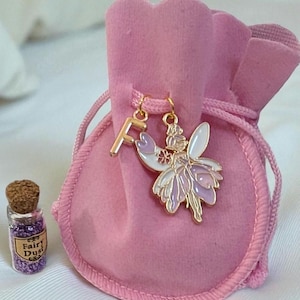 May include: A soft pink velvet drawstring pouch featuring a gold-toned letter F charm and a fairy charm with white and lavender enamel wings. Beside the pouch sits a small glass vial with a cork stopper containing purple glitter labelled Fairy Dust.