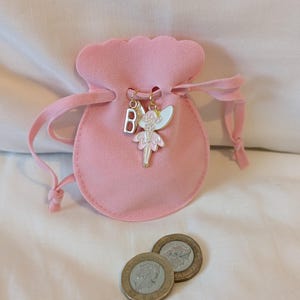 May include: A soft pink velvet drawstring pouch featuring a gold-toned metal charm of a fairy with white and pink enamel wings and dress, alongside a silver-toned metal letter B charm. Two circular metallic coins are positioned in front of the pouch.