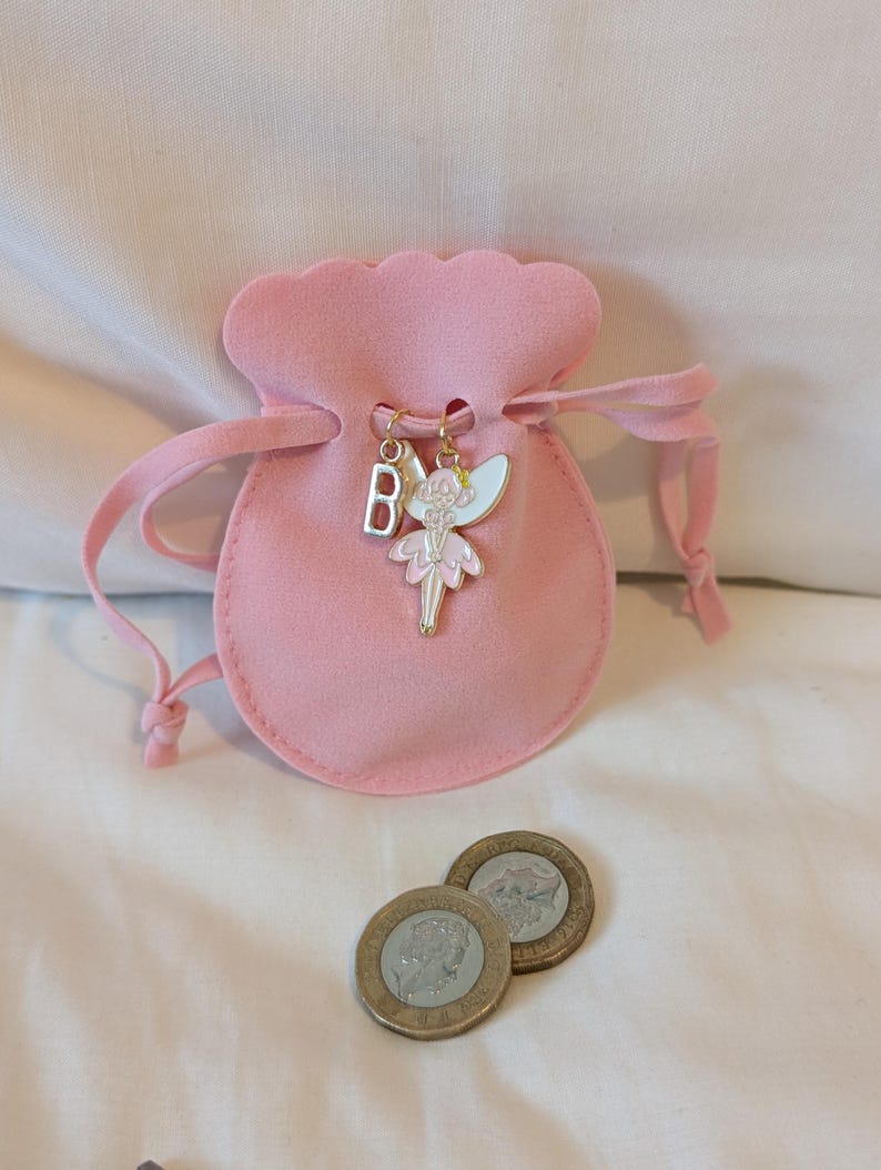 May include: A soft pink velvet drawstring pouch featuring a gold-toned metal charm of a fairy with white and pink enamel wings and dress, alongside a silver-toned metal letter B charm. Two circular metallic coins are positioned in front of the pouch.