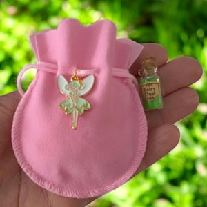 May include: A light pink drawstring pouch featuring a gold-toned metal fairy charm with white and light green enamel wings. Beside the pouch is a small clear glass bottle with a cork stopper, containing green powder and a label reading Fairy Dust.