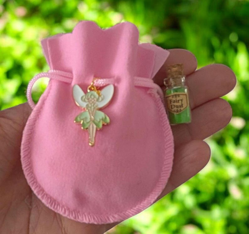 May include: A light pink drawstring pouch featuring a gold-toned metal fairy charm with white and light green enamel wings. Beside the pouch is a small clear glass bottle with a cork stopper, containing green powder and a label reading Fairy Dust.