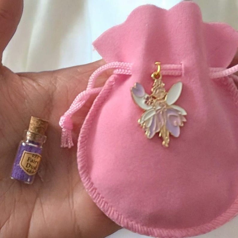 May include: A small pink drawstring pouch featuring a gold-toned metal fairy charm with white and lavender enamel wings. Beside the pouch is a miniature clear glass bottle with a cork stopper, filled with purple glitter labelled Fairy Dust.
