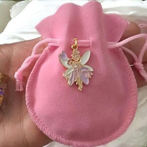 May include: A soft pink velvet drawstring pouch featuring a gold-toned metal fairy pendant with white and lavender enamel wings. Beside the pouch sits a miniature clear glass bottle with a cork stopper, filled with purple glitter labelled Fairy Dust.