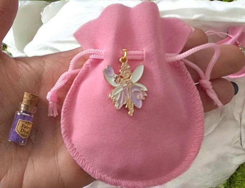 May include: A soft pink velvet drawstring pouch featuring a gold-toned metal fairy pendant with white and lavender enamel wings. Beside the pouch sits a miniature clear glass bottle with a cork stopper, filled with purple glitter labelled Fairy Dust.
