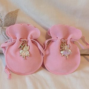 May include: Two light pink velvet drawstring pouches with scalloped edges. Each pouch features a gold-toned metal charm attached to the drawstring, consisting of a letter charm, H on the left and S on the right, paired with a small fairy figure.