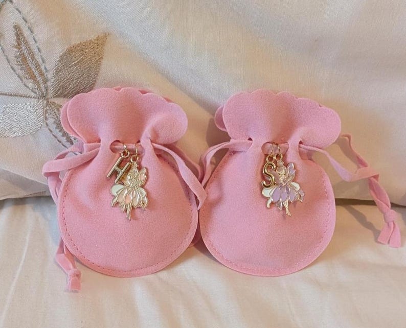 May include: Two light pink velvet drawstring pouches with scalloped edges. Each pouch features a gold-toned metal charm attached to the drawstring, consisting of a letter charm, H on the left and S on the right, paired with a small fairy figure.
