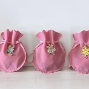 May include: Five small, round, pink fabric drawstring pouches arranged in a row. Each pouch features a different metal fairy charm attached to the front, including figures with white, blue, cream, yellow, and green accents on their wings and clothing.