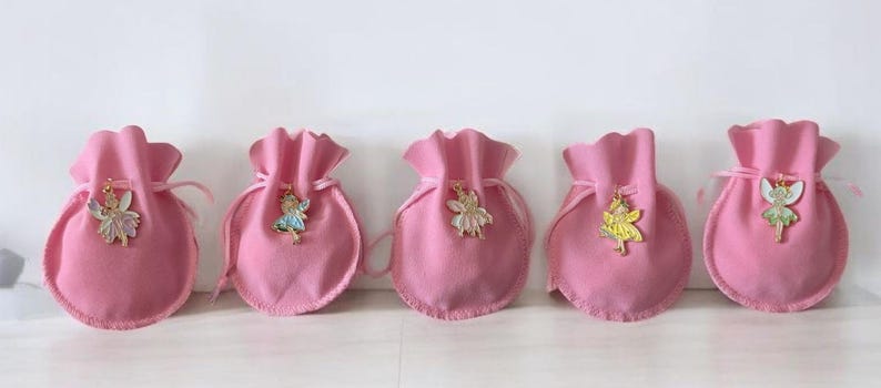May include: Five small, round, pink fabric drawstring pouches arranged in a row. Each pouch features a different metal fairy charm attached to the front, including figures with white, blue, cream, yellow, and green accents on their wings and clothing.