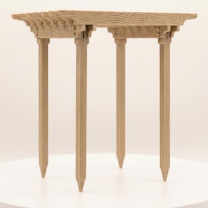 May include: A miniature wooden pergola with intricate wood grain and traditional joinery. The light-colored structure has a rectangular top supported by four slender, pointed legs. The pergola is displayed on a white surface.