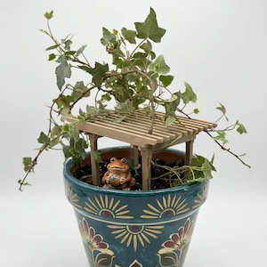 May include: A teal ceramic flower pot with a floral design, holding a small wooden pergola and a frog figurine. Green ivy spills over the top. The pot's design includes sunburst and floral patterns in red, yellow, and cream.