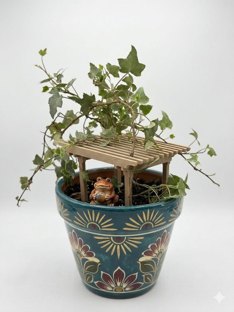 May include: A teal ceramic flower pot with a floral design, holding a small wooden pergola and a frog figurine. Green ivy spills over the top. The pot's design includes sunburst and floral patterns in red, yellow, and cream.