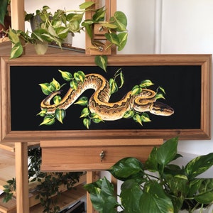 May include: A framed painting of a snake with a yellow and brown pattern, curled around green leaves on a black background.