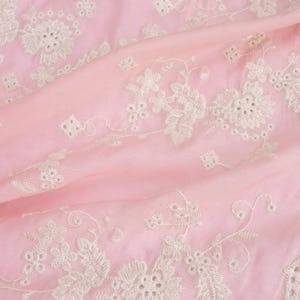 Op de afbeelding: Roze stof met een wit geborduurd bloemenpatroon. De stof heeft een delicate, kant-achtige rand.