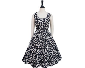 May include: A sleeveless black dress with a white daisy pattern. The dress has a fitted bodice and a flared skirt, creating a classic vintage silhouette. The dress is displayed on a mannequin against a white background.