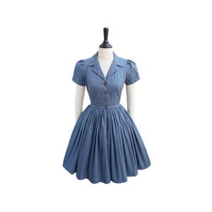 Könnte beinhalten: Ein blaues Baumwollkleid mit Kragen, kurzen Ärmeln und einer Knopfleiste vorne. Das Kleid hat einen weiten Rock und ist an der Taille tailliert.