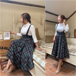 May include: A woman wearing a white turtleneck top with a green and black plaid pinafore dress. The dress has adjustable straps and a flared skirt. She is standing on a tiled floor.
