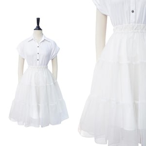 May include: A white, layered tulle skirt with a white button-up shirt. The skirt has a gathered waistband and is knee-length.