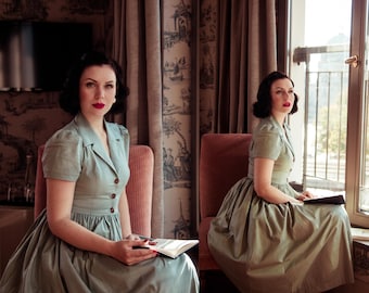 May include: Two women in vintage-style dresses sit in chairs in a room with large windows. They are both reading books and have similar hairstyles. The room has a patterned wallpaper and a dark wood floor.