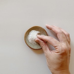 May include: A small, round, beige ceramic dish with a speckled texture and a dark brown rim. The dish contains a small pile of white, crystalline granules. The object is circular in shape and features a smooth, slightly concave surface.