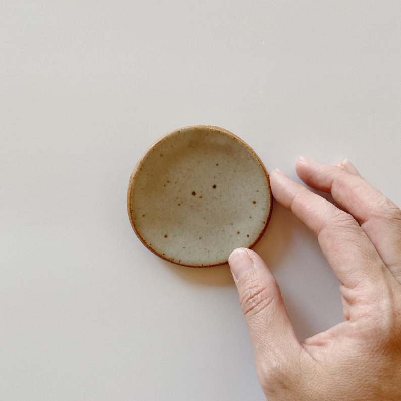 May include: A small, round ceramic dish with a speckled light gray glaze and a contrasting dark brown, unglazed rim. The dish has a slightly irregular, organic shape and a smooth, concave center, showcasing a handcrafted appearance.
