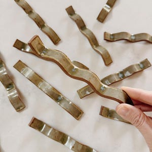 May include: A collection of ceramic wavy-shaped handles in shades of brown and beige. The handles have a textured, glazed finish and are arranged on a light-colored surface. One handle is being held by a hand.