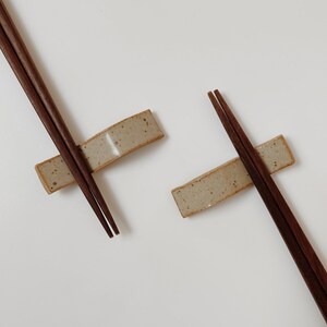 May include: Two pairs of brown wooden chopsticks resting on two rectangular ceramic chopstick holders. The holders are a light beige color with a speckled texture.