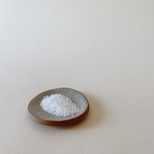 May include: A small, round, shallow ceramic dish with a speckled grey interior and a raw, unglazed brown clay rim. A mound of white, crystalline granular substance is centered on the dish, contrasting with the smooth, muted grey surface.