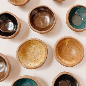 May include: A selection of small, handmade ceramic bowls in various earth tones. The bowls have a rustic, organic shape with a textured exterior and glazed interiors in shades of brown, yellow, and teal. The bowls are arranged on a white surface.