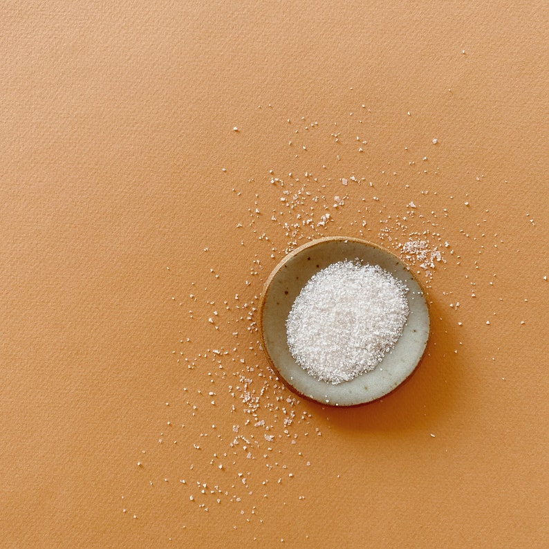 May include: A small, round, light-colored ceramic dish filled with a mound of white crystalline grains. Additional white grains are scattered around the dish, creating a textured appearance with a contrast between the white and the tan surface.