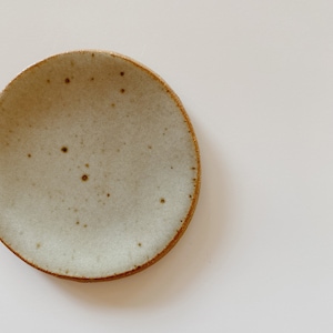 May include: A circular ceramic plate with a speckled off-white glaze and a visible brown clay rim. The surface features small, dark brown spots distributed across the center, while the edges show the natural texture of the unglazed clay base.