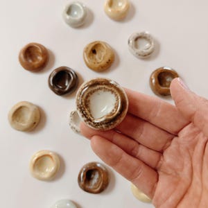 Puede incluir: Una colección de pequeños cuencos redondos de cerámica en varios colores tierra. Uno de los cuencos se sostiene en una mano, mostrando un interior blanco con un borde moteado marrón. Otros cuencos están dispersos sobre una superficie blanca.