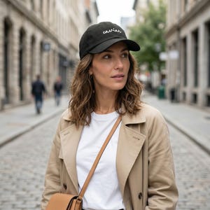 Pode incluir: Bon&eacute; de beisebol preto com a palavra "caviar" bordada em branco. O bon&eacute; tem uma aba curva. A pessoa est&aacute; usando uma camiseta branca e um trench coat bege. Uma bolsa de ombro de couro marrom tamb&eacute;m &eacute; vis&iacute;vel.