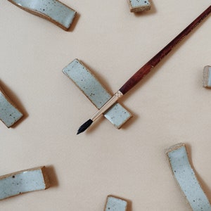 Peut inclure: Un pinceau de peinture à l'aquarelle repose sur un repose-pinceau en céramique bleu clair. Le repose-pinceau est rectangulaire avec des détails mouchetés et un bord marron. Le pinceau a un manche en bois foncé et une virole argentée. Le fond est beige uni.