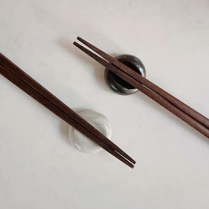 May include: Two pairs of dark brown wooden chopsticks rest on small, round chopstick rests. One rest is a speckled white, the other is a dark, glossy brown. The chopsticks are angled across a white surface.