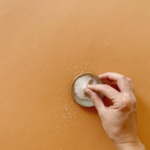 May include: A small, round, light-colored ceramic dish containing a mound of white granulated sugar. A portion of the sugar is being pinched and lifted from the dish, with scattered grains of sugar resting on the surrounding surface area.