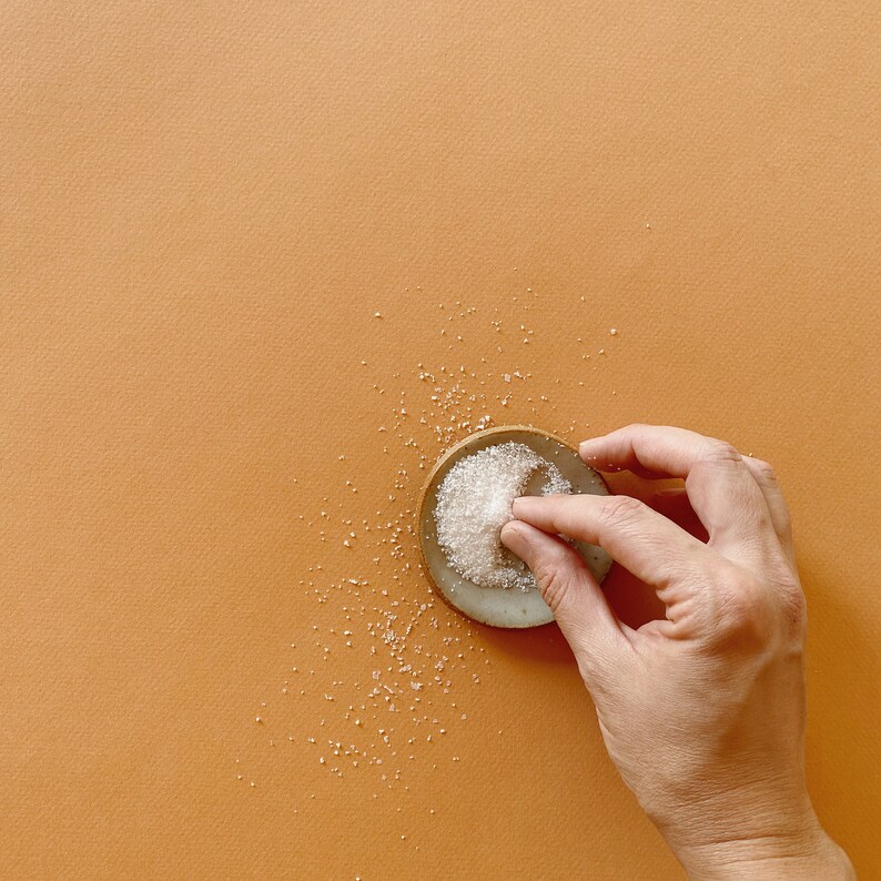 May include: A small, round, light-colored ceramic dish containing a mound of white granulated sugar. A portion of the sugar is being pinched and lifted from the dish, with scattered grains of sugar resting on the surrounding surface area.