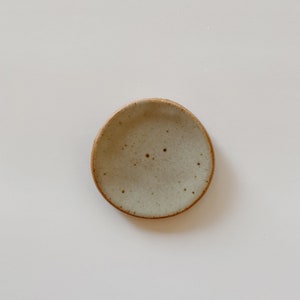 May include: A small, round ceramic dish with a speckled light gray glaze and a visible brown clay rim. The surface of the dish features a subtle, uneven texture with dark brown flecks distributed across the interior, creating a natural appearance.