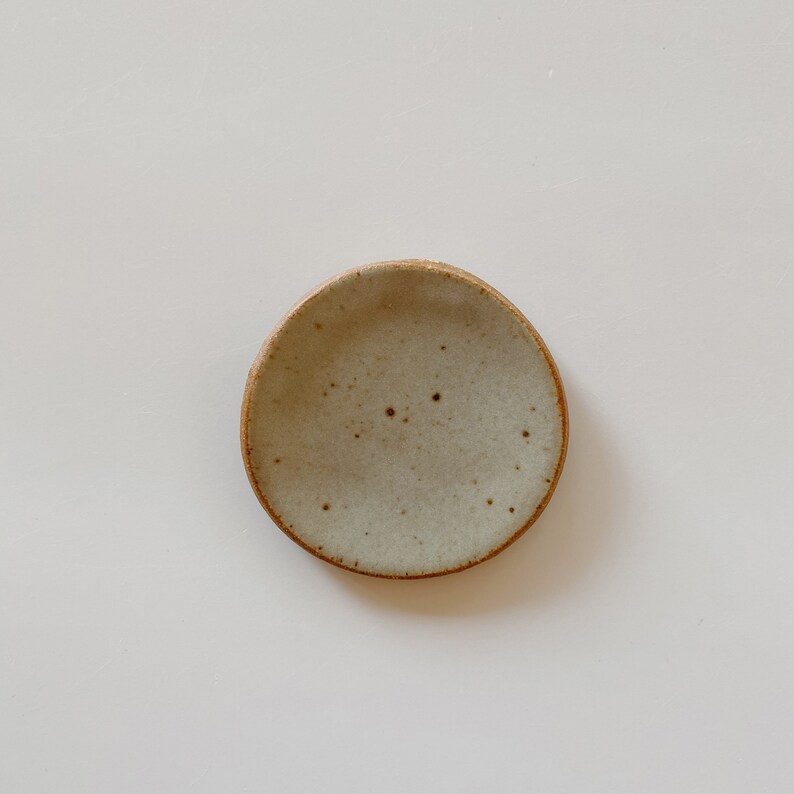 May include: A small, round ceramic dish with a speckled light gray glaze and a visible brown clay rim. The surface of the dish features a subtle, uneven texture with dark brown flecks distributed across the interior, creating a natural appearance.
