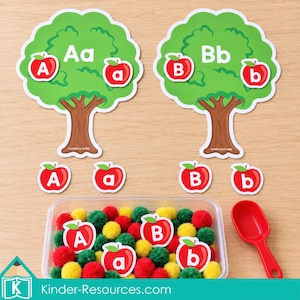 Op de afbeelding: Twee groene boomuitsparingen met het label Aa en Bb met rode appelpictogrammen met de letters A, a, B en b. Inclusief losse rode appeluitsparingen met de letters A, a, B, b, een doorzichtige plastic bak gevuld met pompons en een rode schep.