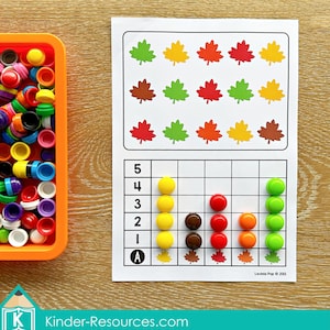 Könnte beinhalten: Ein Lernspielset mit einem bedruckten Blatt mit bunten Blätterillustrationen und einem Zählgitter. Enthält einen orangefarbenen Behälter mit verschiedenfarbigen Zählern. Das Blatt trägt den Text "Kinder-Resources.com".