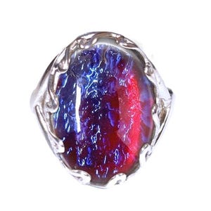 May include: A silver ring with an oval-shaped, iridescent stone. The stone features a blend of red and blue hues, creating a vibrant, eye-catching effect. The ring's setting has a decorative, openwork design. The ring is set against a white background.