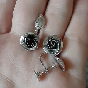 May include: A pair of silver rose-shaped clip-on earrings with a leaf detail. The earrings have a screw-back closure.