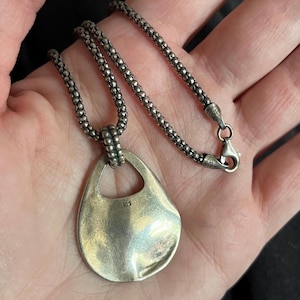 May include: A sterling silver necklace featuring a textured, beaded chain and a smooth, teardrop-shaped pendant. The pendant has a cutout at the top and is stamped with 925, indicating sterling silver composition. The chain includes a lobster clasp.