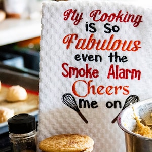 May include: White kitchen towel with red and orange embroidery that reads "My Cooking is so Fabulous even the Smoke Alarm Cheers me on".