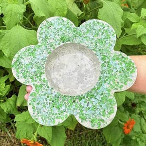 May include: A flower-shaped concrete dish featuring a central circular basin surrounded by six rounded petals. The surface of the petals is embedded with numerous small, translucent green and light blue glass pebbles, creating a textured finish.