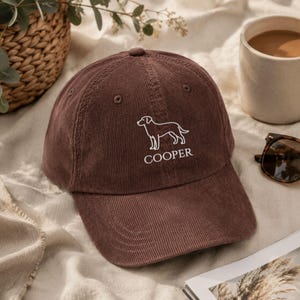 May include: A brown corduroy baseball cap featuring a white embroidered outline of a dog and the text COOPER centred below the graphic. The cap has a structured crown, a curved brim, and matching brown eyelets on the top panels.