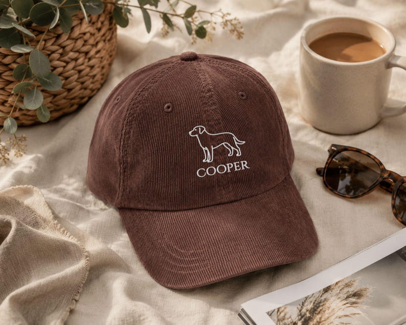 May include: A brown corduroy baseball cap featuring a white embroidered outline of a dog and the text COOPER centred below the graphic. The cap has a structured crown, a curved brim, and matching brown eyelets on the top panels.