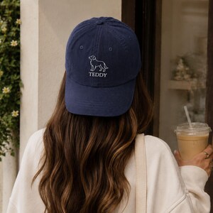 May include: A navy blue baseball cap featuring an embroidered white line art illustration of a dog above the text TEDDY in a simple white font. The cap has a structured crown, a curved brim, and a button at the top centre of the fabric panels.